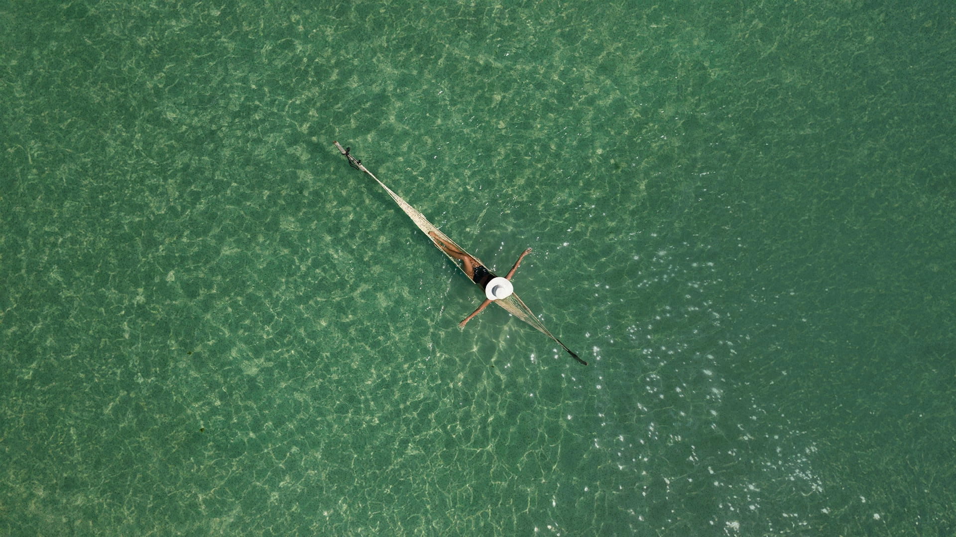Imagem aérea de uma mulher descansando em uma rede instalada sobre as águas rasas e cristalinas de um mar verde-esmeralda da Praia de Patacho. A transparência da água revela texturas de areia e corais no fundo, enquanto o sol cria reflexos cintilantes na superfície, transmitindo uma paz absoluta e inigualável.