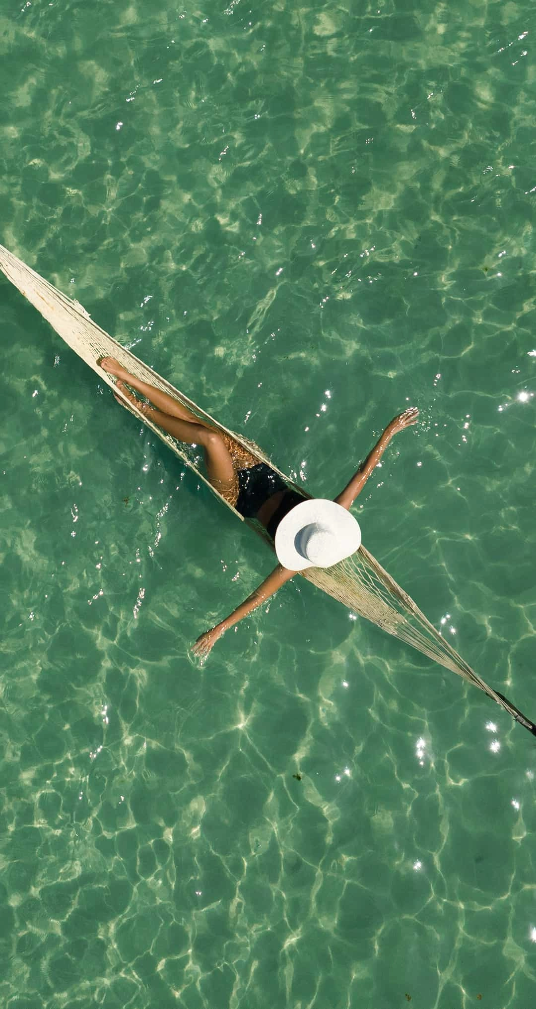  Imagem aérea de uma mulher descansando em uma rede instalada sobre as águas rasas e cristalinas de um mar verde-esmeralda da Praia de Patacho. A transparência da água revela texturas de areia e corais no fundo, enquanto o sol cria reflexos cintilantes na superfície, transmitindo uma paz absoluta e inigualável.