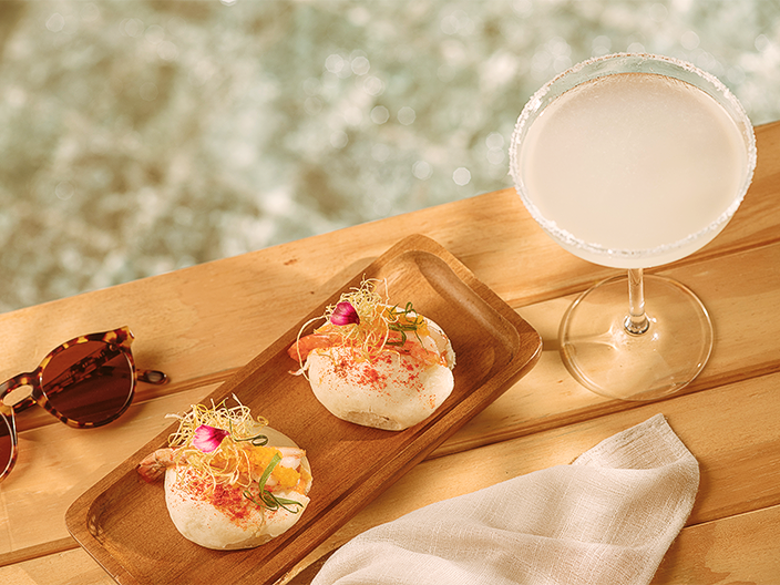 Tábua de madeira servindo dois bao buns recheados com camarões grelhados e finalizados com flores e brotos. Ao lado, um coquetel refrescante em taça coupe com borda de sal e óculos de sol tartaruga, dispostos em um deck de madeira sob a luz solar suave, com o brilho da água da piscina ao fundo.