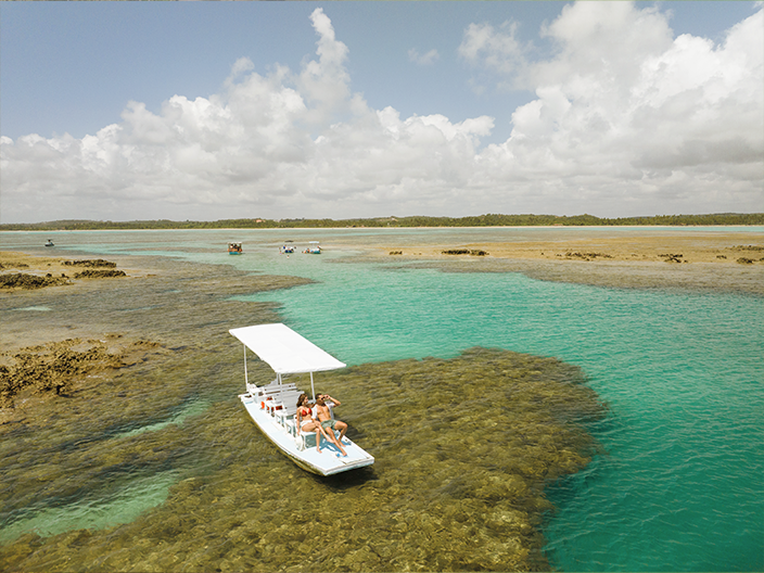 Casal relaxando em um passeio de barco sobre as piscinas naturais da Praia de Patacho, com tons de azul e verde, em meio a recifes e céu aberto.