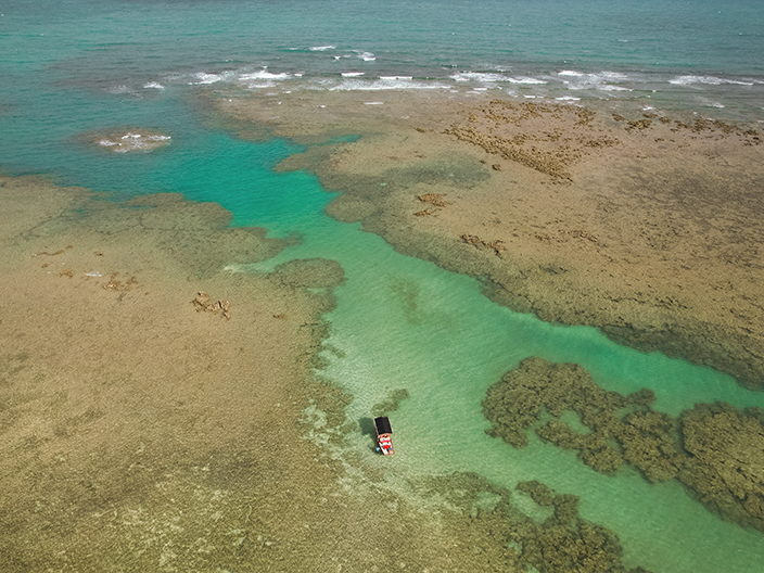 Imagem aérea de um barco de passeio navegando por um canal de águas cristalinas e turquesas, cercado por vastas formações de recifes de corais em tons de marrom e dourado.
